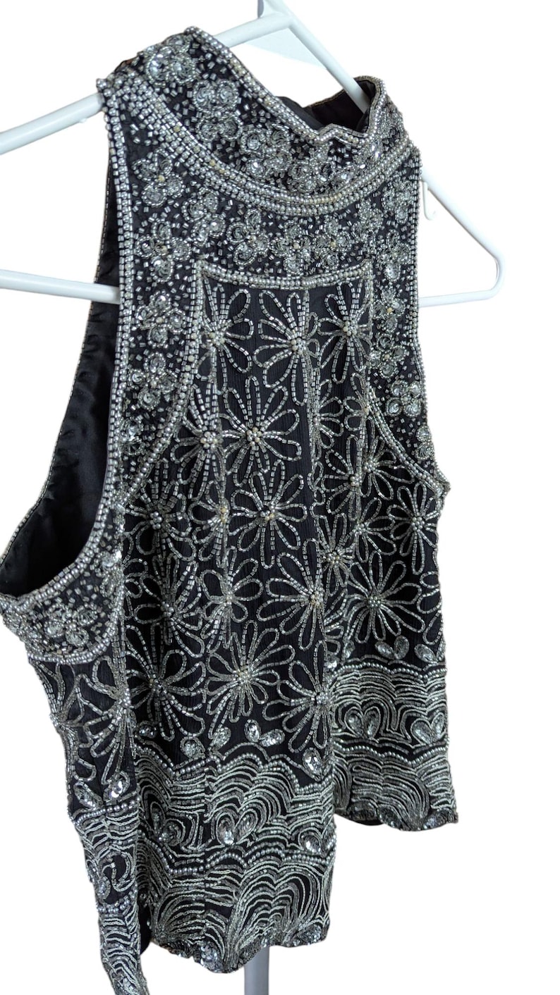 May include: Black sleeveless top with intricate silver bead and sequin floral and wave patterns. The top is embellished with sparkling beads and sequins, creating a glamorous and eye-catching design. The garment is hanging on a white hanger.