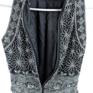 May include: Black sleeveless vest with intricate silver bead and sequin embellishments. The vest features a floral design and a scalloped hem. The zipper is open, revealing the black lining. The garment is hanging on a white hanger.