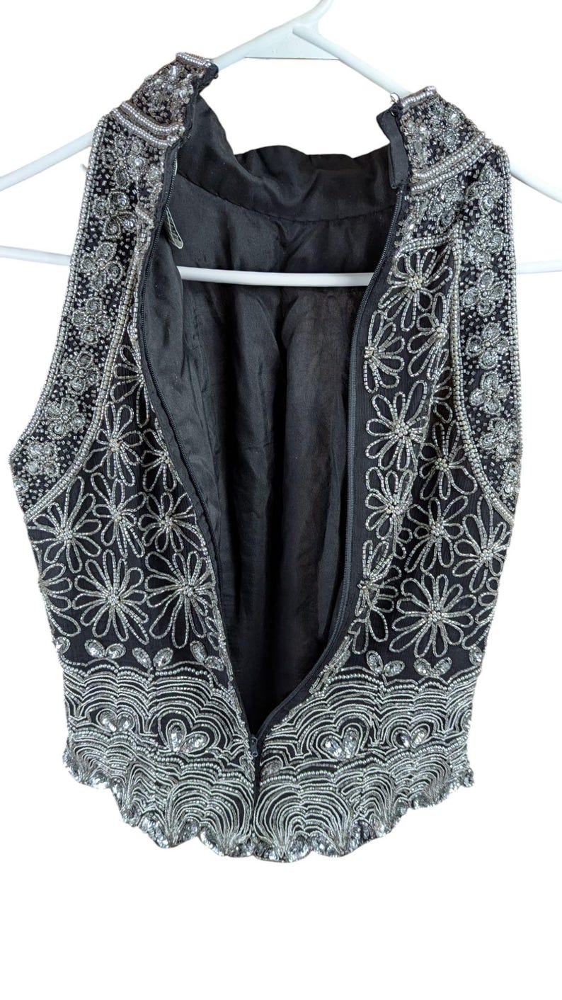 May include: Black sleeveless vest with intricate silver bead and sequin embellishments. The vest features a floral design and a scalloped hem. The zipper is open, revealing the black lining. The garment is hanging on a white hanger.