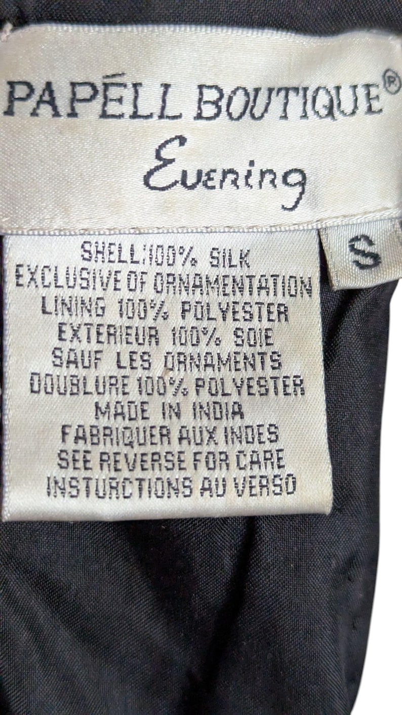 May include: A close-up of a Papell Boutique Evening label. The label is white with black text, including "SHELL: 100% SILK" and "MADE IN INDIA". The size is "S". The background is black.