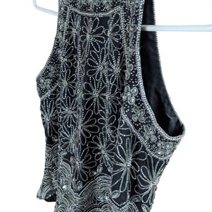 May include: Black sleeveless top with intricate silver bead and sequin embellishments. The top features a high neckline and a floral pattern design. The garment is hanging on a white hanger.