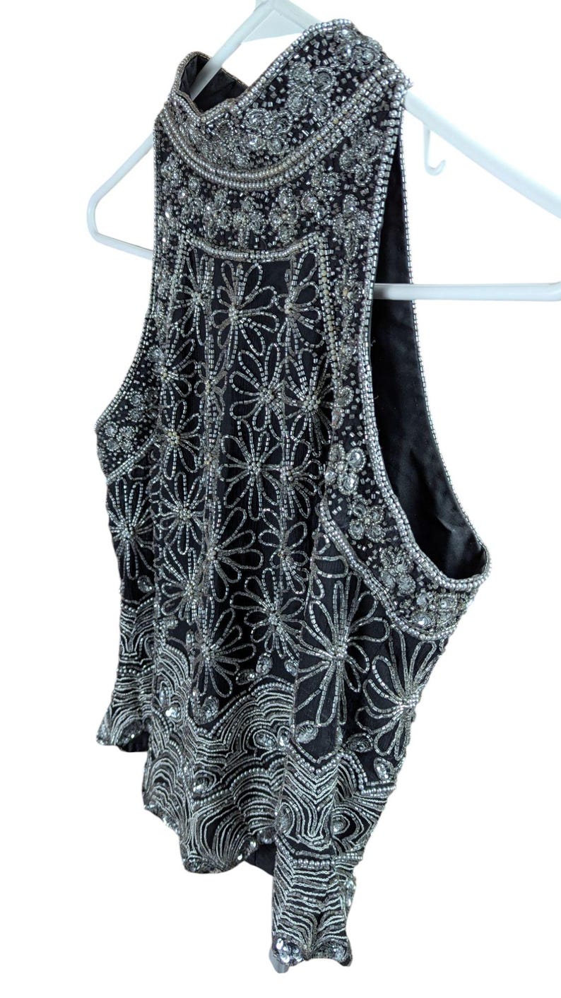 May include: Black sleeveless top with intricate silver bead and sequin embellishments. The top features a high neckline and a floral pattern design. The garment is hanging on a white hanger.