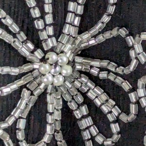 May include: Close-up of a decorative floral design. The design is made of silver-colored beads and small, round, pearl-like beads. The beads are arranged on a dark background, creating a textured pattern.