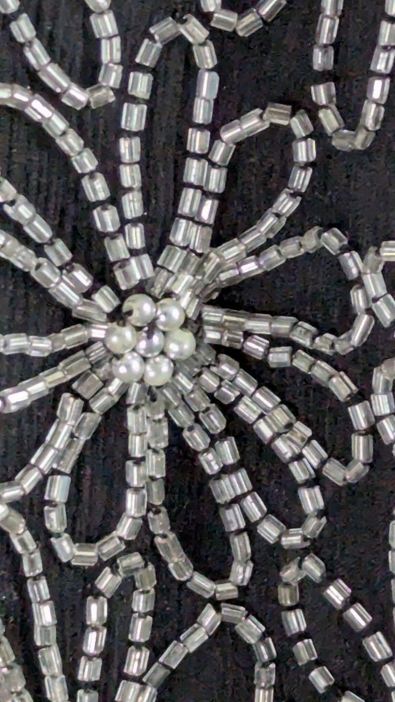 May include: Close-up of a decorative floral design. The design is made of silver-colored beads and small, round, pearl-like beads. The beads are arranged on a dark background, creating a textured pattern.