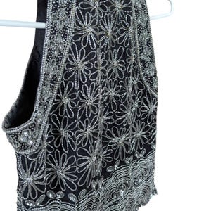 May include: Black sleeveless top with intricate silver bead and sequin floral design. The top is embellished with silver beads and sequins, creating a shimmering effect. The garment is hanging on a white hanger.