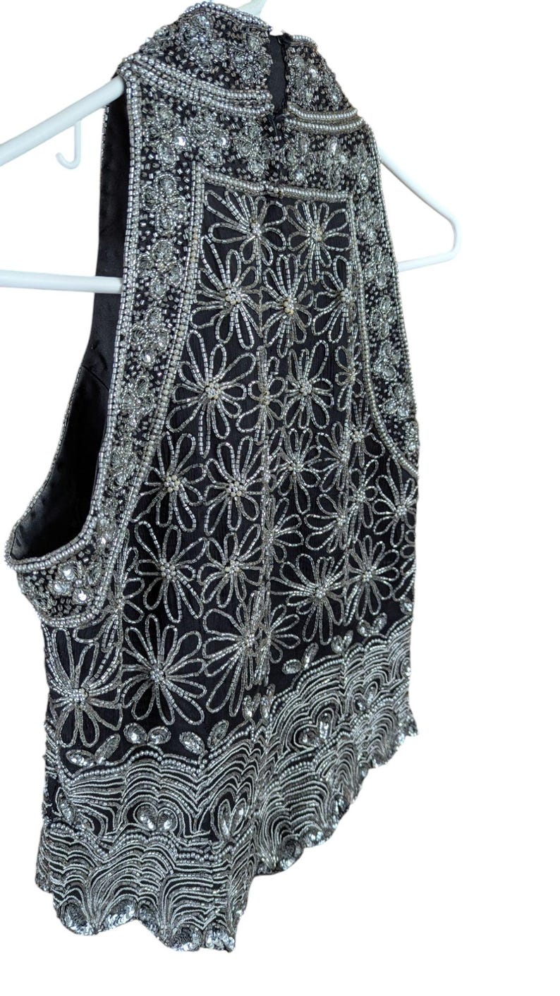 May include: Black sleeveless top with intricate silver bead and sequin floral design. The top is embellished with silver beads and sequins, creating a shimmering effect. The garment is hanging on a white hanger.