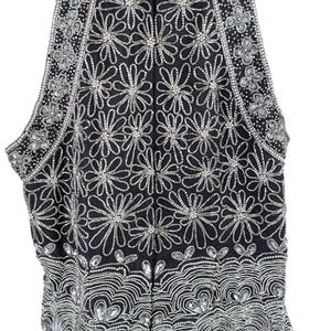 May include: Black sleeveless vest heavily embellished with silver sequins and beads. The vest features a floral pattern throughout, with a scalloped hemline. The neckline and armholes are also decorated with silver embellishments. A zipper runs down the front.