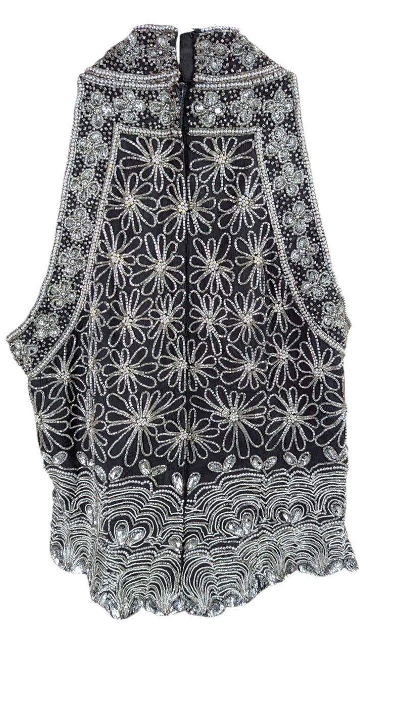 May include: Black sleeveless vest heavily embellished with silver sequins and beads. The vest features a floral pattern throughout, with a scalloped hemline. The neckline and armholes are also decorated with silver embellishments. A zipper runs down the front.
