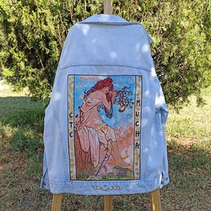 Peut inclure: Une veste en jean bleu clair avec une peinture d'une femme en robe fluide au dos. La peinture est dans un cadre doré et comprend le texte "Mucha" et "Été."