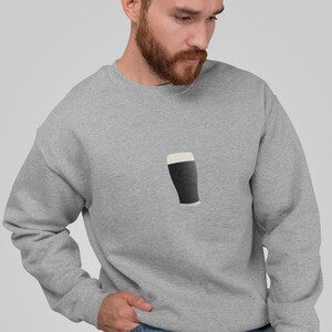 May include: A light gray sweatshirt featuring a graphic of a pint of stout. The pint is dark with a white foamy head. The sweatshirt is made of a soft material.