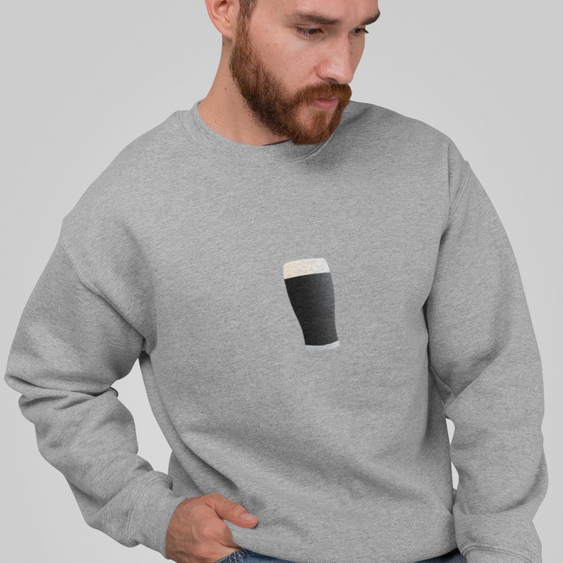 May include: A light gray sweatshirt featuring a graphic of a pint of stout. The pint is dark with a white foamy head. The sweatshirt is made of a soft material.