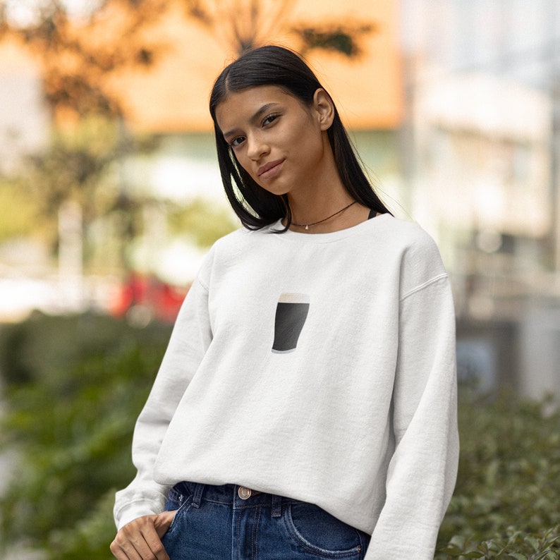May include: White sweatshirt featuring a graphic of a pint of stout. The design is a simple illustration of a dark beer with a creamy head. The sweatshirt is a casual, everyday garment, suitable for various occasions.