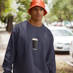 May include: Navy blue sweatshirt featuring a graphic of a pint of stout. The sweatshirt has a crew neck and long sleeves. The person in the image is wearing an orange bucket hat. The background is blurred, suggesting an outdoor setting.