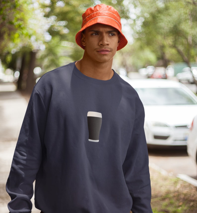 May include: Navy blue sweatshirt featuring a graphic of a pint of stout. The sweatshirt has a crew neck and long sleeves. The person in the image is wearing an orange bucket hat. The background is blurred, suggesting an outdoor setting.