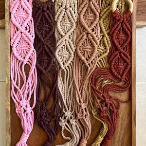 以下が含まれることがあります： ピンク、ブラウン、ベージュ、イエロー、レッドなど、さまざまな色のマクラメ製のプラントハンガーが6つあります。各ハンガーは、上部に木製リング、下部にフリンジが付いています。