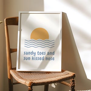 Może przedstawiać: Biały wydruk w ramie z żółtym słońcem zachodzącym nad niebieskimi falami. Tekst "sandy toes and sun kissed nose" jest napisany na niebiesko pod obrazem.