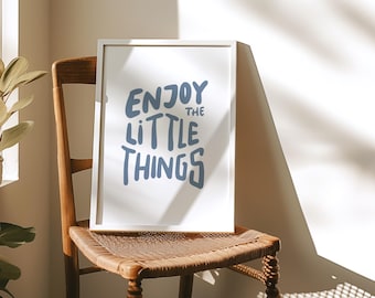 Puede incluir: Una impresión enmarcada en blanco con el texto "Enjoy the little things" en letras azules sobre un fondo blanco. La impresión está sentada en una silla de madera.