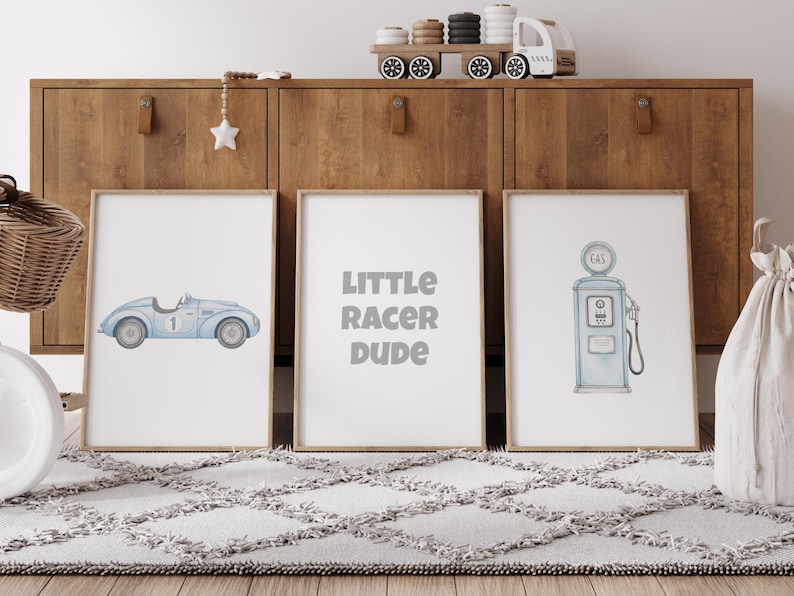 May include: Three framed prints with a racing theme. One features a light blue race car, another reads "LITTLE RACER DUDE," and the third depicts a vintage gas pump, all in soft watercolor style. The prints are displayed on a rug.