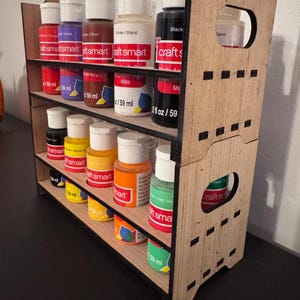 May include: A wooden paint bottle organiser with two shelves. The organiser is made of light brown wood with black trim. It holds 10 bottles of acrylic paint in various colours, including red, purple, brown, white, black, yellow, orange, and green. The bottles are labelled "craft smart" and have a capacity of 59 ml.