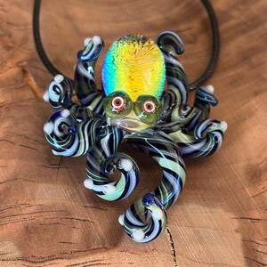 May include: A glass octopus pendant with a black cord. The octopus has a yellow and green iridescent head, green eyes, and striped tentacles in shades of blue, black, and white. The pendant is displayed on a wooden surface.