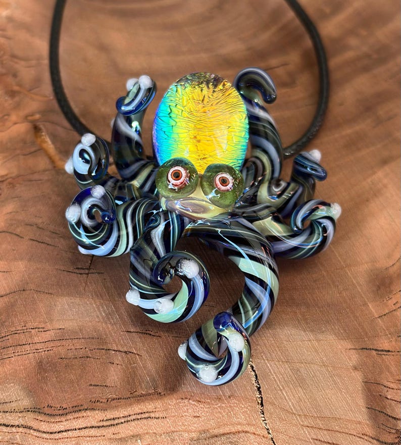 May include: A glass octopus pendant with a black cord. The octopus has a yellow and green iridescent head, green eyes, and striped tentacles in shades of blue, black, and white. The pendant is displayed on a wooden surface.