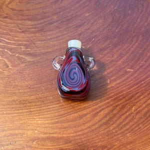 May include: A small, red, black, and white glass bottle with two handles. The bottle has a swirl design.