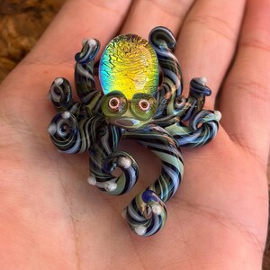 May include: A handcrafted glass octopus figurine with a dichroic glass head that reflects yellow and green hues. The octopus has green, black, and blue striped tentacles with white accents. The eyes are red and the body is green.