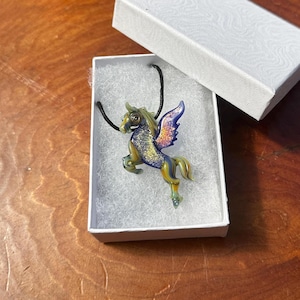 May include: A glass winged horse pendant with iridescent blue and gold wings, on a black cord, in a white box with white tissue paper.
