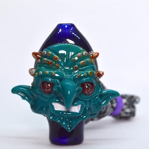 May include: A glass pipe with a green and blue goblin face design. The goblin has red eyes, a wide mouth, and a pointed nose. The pipe is decorated with small orange and white dots.