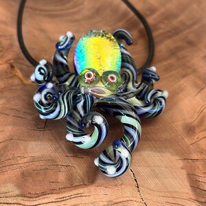 May include: A glass octopus pendant with a black cord. The octopus features a yellow and iridescent head, green eyes, and striped tentacles in shades of blue, black, and white. The pendant is a unique piece of art.