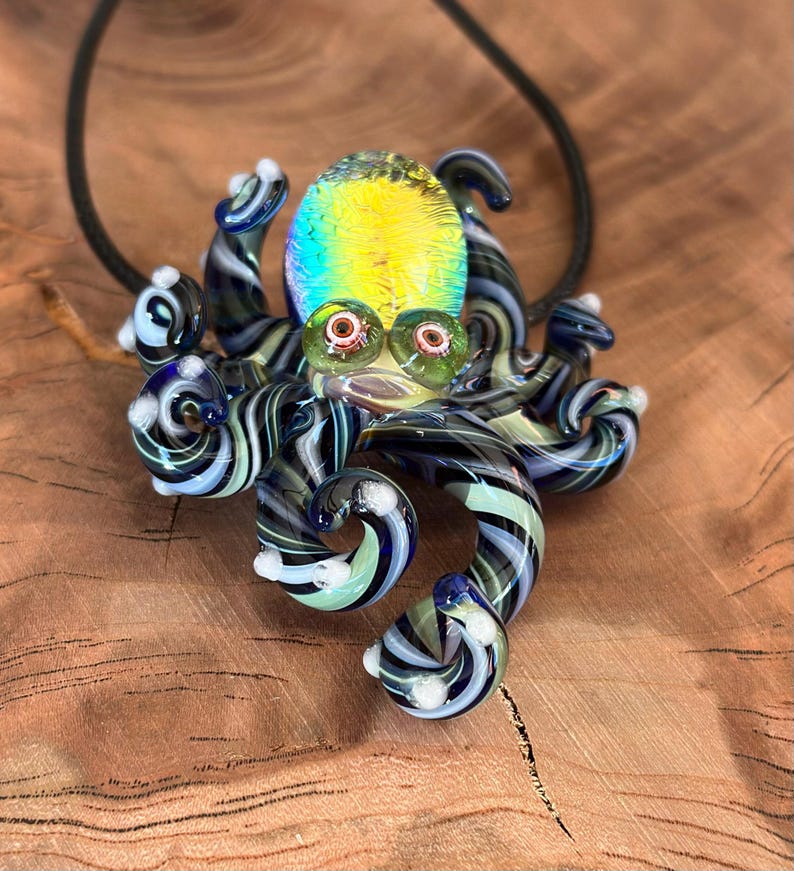 May include: A glass octopus pendant with a black cord. The octopus features a yellow and iridescent head, green eyes, and striped tentacles in shades of blue, black, and white. The pendant is a unique piece of art.