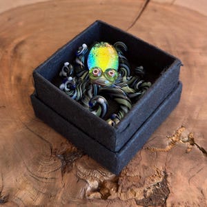 May include: A colorful glass octopus figurine with a rainbow-colored head and red eyes, resting in a black box. The octopus has striped tentacles in shades of black and green. The box is on a wooden surface.
