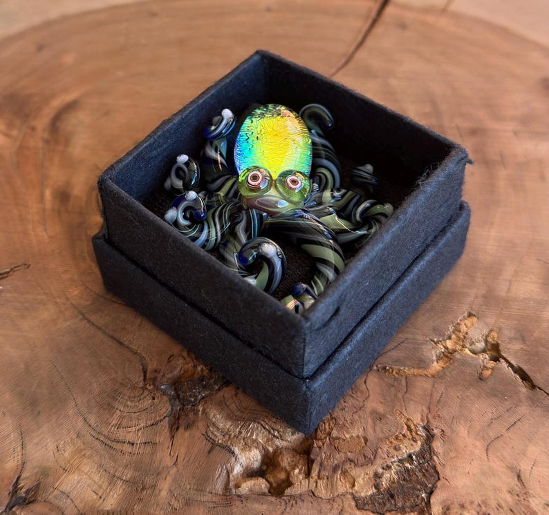 May include: A colorful glass octopus figurine with a rainbow-colored head and red eyes, resting in a black box. The octopus has striped tentacles in shades of black and green. The box is on a wooden surface.