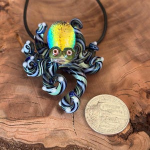 May include: A glass octopus pendant with a black cord. The octopus has a yellow, blue, and green head, with black and white striped tentacles. The pendant is next to a quarter coin.