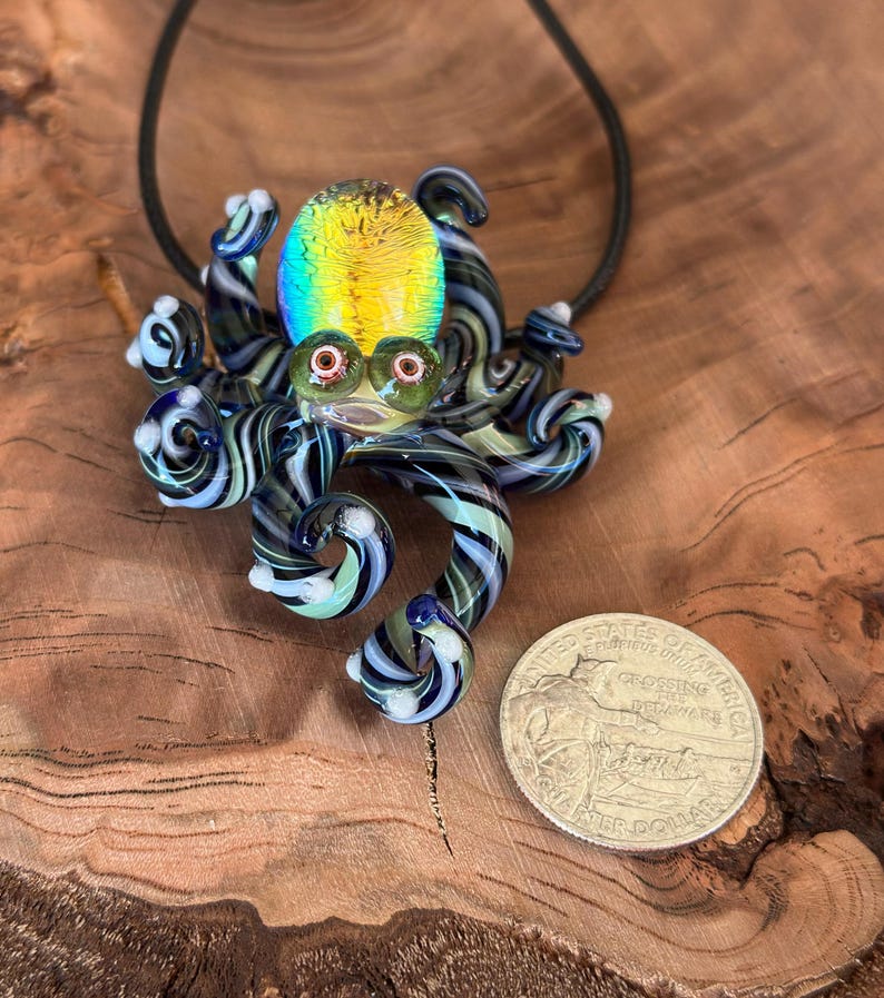 May include: A glass octopus pendant with a black cord. The octopus has a yellow, blue, and green head, with black and white striped tentacles. The pendant is next to a quarter coin.