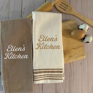 May include: Two kitchen towels, one taupe and one cream, with the embroidered text "Ellen's Kitchen." The cream towel has brown stripes. A wooden cutting board, rolling pin, and spoon are in the background.