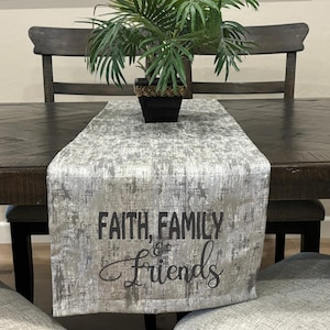 May include: A gray and white table runner with the text "Faith, Family & Friends" printed in black. The runner is on a dark wood table with a green plant in a black pot.