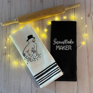 May include: A wooden rolling pin rests above two kitchen towels. One white towel features a snowman design with a black hat and blue snowflakes. The other towel is black with the embroidered text "Snowflake MAKER".