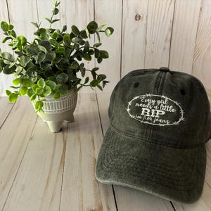 May include: A dark green, distressed baseball cap with white embroidery that reads "Every girl needs a little RIP in her jeans". The cap has a barbed wire design around the text.