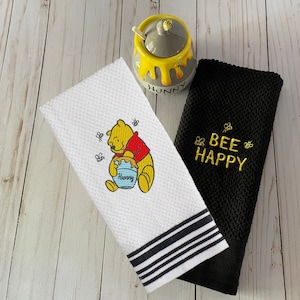 May include: Two kitchen towels, one white with black stripes and a Winnie the Pooh embroidery, the other black with gold embroidery that says "Bee Happy". A yellow honey pot with a gray lid and a bee on top is also in the image.
