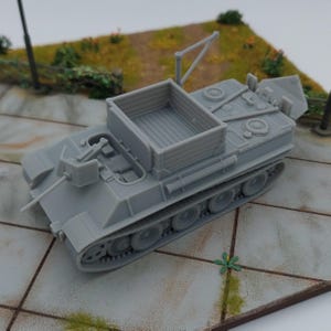 Puede incluir: Un modelo de tanque gris con un diseño detallado, que presenta un área de carga rectangular, un cañón y orugas visibles. El tanque en miniatura está sobre una base de diorama con una superficie pavimentada y vegetación.