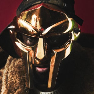 May include: A person wearing a gold-colored mask with a sharp, angular design. The mask covers the person's entire face and has a menacing expression. The person is wearing a brown fur-lined jacket.
