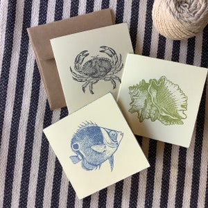 May include: Three blank greeting cards with illustrations of a crab, a seashell, and a fish. The cards are on a striped background.