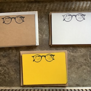 May include: Three stacks of note cards with a black and white illustration of a pair of glasses on the front. The stacks are brown, white, and yellow.