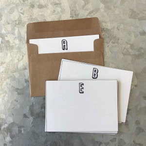 May include: Three white note cards with black letters on them. The letters are A, B, and E. The cards are stacked on top of each other and are sitting on a brown envelope.