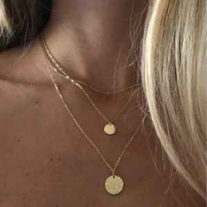 K&ouml;nnte beinhalten: Ein mehrschichtiges goldenes Halsketten-Set. Die Halsketten haben zarte Ketten mit kleinen, runden Anh&auml;ngern. Der unterste Anh&auml;nger hat eine geh&auml;mmerten Textur, w&auml;hrend die anderen glatt sind. Der Schmuck wird auf der Haut einer Person pr&auml;sentiert.
