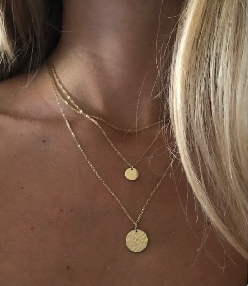 K&ouml;nnte beinhalten: Ein mehrschichtiges goldenes Halsketten-Set. Die Halsketten haben zarte Ketten mit kleinen, runden Anh&auml;ngern. Der unterste Anh&auml;nger hat eine geh&auml;mmerten Textur, w&auml;hrend die anderen glatt sind. Der Schmuck wird auf der Haut einer Person pr&auml;sentiert.