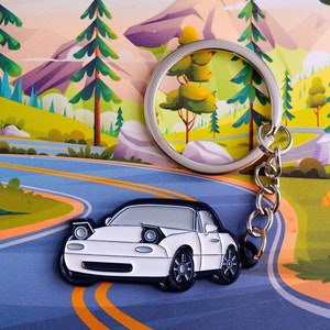 Peut inclure: Porte-clés en métal blanc en forme de voiture de sport avec un anneau et une chaîne argentés. Le porte-clés est sur une route sinueuse bleue et orange avec des arbres verts et une montagne en arrière-plan.