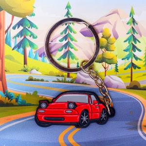 May include: A red keychain car charm with a silver key ring. The car is a small, sporty model with a black interior and black wheels. The keychain is set against a cartoon background of a winding road and trees.