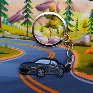 May include: A silver keychain with a black car charm. The car charm is a detailed model of a sports car with a black finish. The keychain is hanging on a winding road with a mountain in the background.